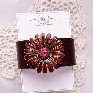 Puede incluir: Una pulsera de cuero marr&oacute;n oscuro con un adorno met&aacute;lico grande en forma de margarita, con p&eacute;talos de color rojo intenso y un centro de pedrer&iacute;a rosa brillante. La pulsera se presenta sobre una tarjeta blanca con el texto Klay Kreations Handmade Jewelry.