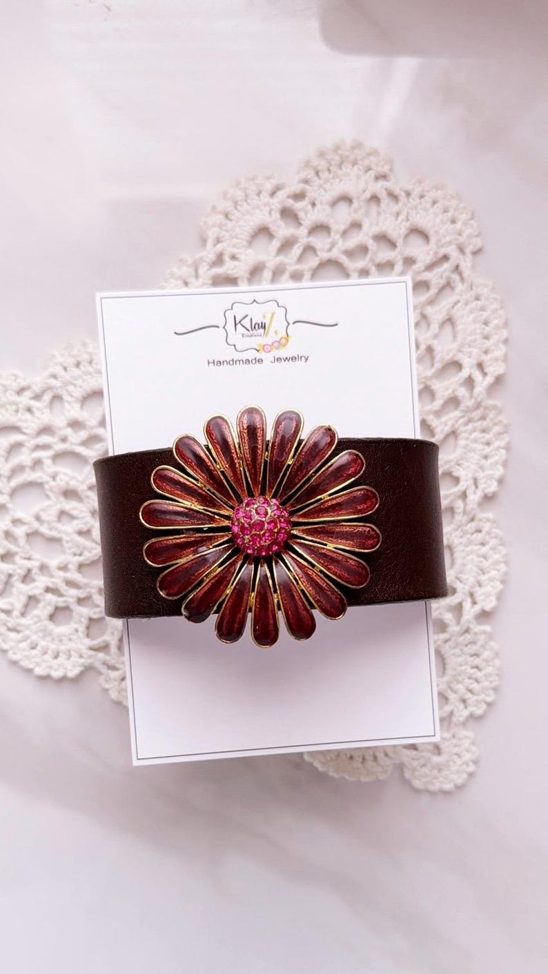 Puede incluir: Una pulsera de cuero marr&oacute;n oscuro con un adorno met&aacute;lico grande en forma de margarita, con p&eacute;talos de color rojo intenso y un centro de pedrer&iacute;a rosa brillante. La pulsera se presenta sobre una tarjeta blanca con el texto Klay Kreations Handmade Jewelry.