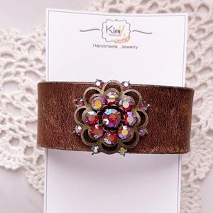 Pu&ograve; includere: Un bracciale a fascia in pelle marrone con un ornamento centrale circolare in filigrana di ottone anticato, decorato con cristalli sfaccettati iridescenti rosa e rossi. Il bracciale &egrave; esposto su un cartoncino bianco con la scritta Klay Handmade Jewelry.
