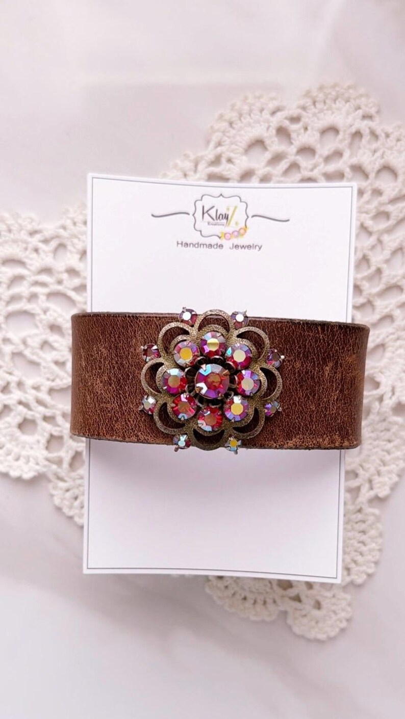 Pu&ograve; includere: Un bracciale a fascia in pelle marrone con un ornamento centrale circolare in filigrana di ottone anticato, decorato con cristalli sfaccettati iridescenti rosa e rossi. Il bracciale &egrave; esposto su un cartoncino bianco con la scritta Klay Handmade Jewelry.