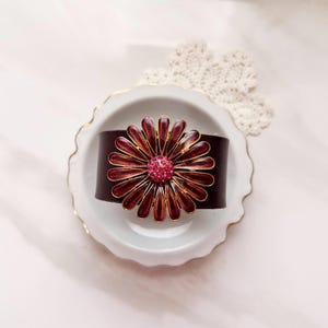 Puede incluir: Una pulsera de cuero marr&oacute;n oscuro con un adorno floral met&aacute;lico grande. La flor tiene p&eacute;talos de esmalte rojo intenso con bordes dorados y un centro de peque&ntilde;os brillantes de color rosa intenso, creando un punto focal floral llamativo.