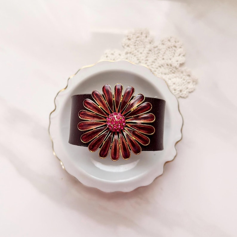 Puede incluir: Una pulsera de cuero marr&oacute;n oscuro con un adorno floral met&aacute;lico grande. La flor tiene p&eacute;talos de esmalte rojo intenso con bordes dorados y un centro de peque&ntilde;os brillantes de color rosa intenso, creando un punto focal floral llamativo.