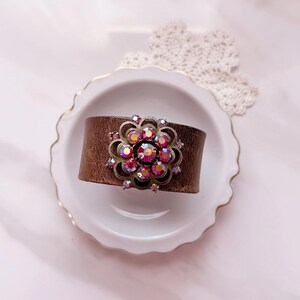 Pu&ograve; includere: Un ampio bracciale a fascia in pelle marrone con un ornamento centrale circolare in metallo. L'ornamento ha la forma di un fiore stilizzato con dettagli in filigrana ed &egrave; incastonato con molteplici cristalli sfaccettati rosa e rossi.