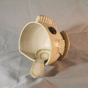 May include: A ceramic bowl shaped like a fish with a small spoon inside. The bowl is white with a light green glaze and has a black eye. The spoon is also white with a light green glaze.