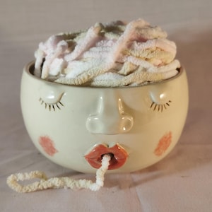 May include: A cream-coloured ceramic yarn bowl shaped like a face with closed eyes, a nose, and pink cheeks. The bowl features red lips with a small hole in the centre, through which a strand of soft, variegated white and pale pink yarn is threaded.