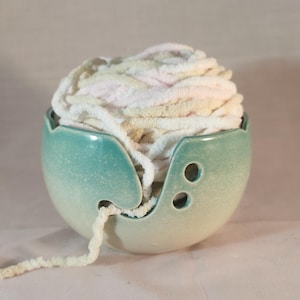 Puede incluir: Un cuenco de cerámica para lana con un esmalte degradado en color verde azulado y crema. El cuenco presenta un corte en espiral para guiar el hilo y dos pequeños orificios circulares para agujas de tejer. Un ovillo de lana suave en tonos crema y rosa.