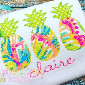 May include: Three embroidered pineapple designs in bright colors with white polka dots on a white fabric background. The word "claire" is embroidered in pink below the pineapples.