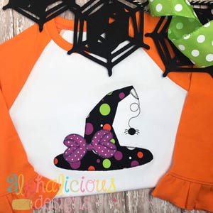 May include: A white and orange long-sleeved shirt with a black and white witch hat with a purple bow and polka dots. The hat has a spider hanging from it. The shirt has the text "alphadelicious designs" on the bottom.