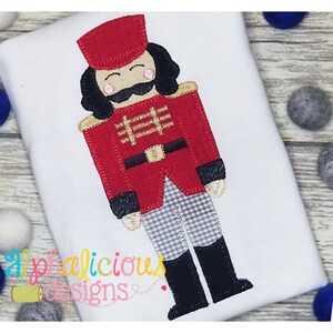 May include: A red and black felt applique of a toy soldier with a black and white checkered pattern on the pants. The soldier has a red hat, a black mustache, and rosy cheeks.