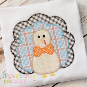 May include: Embroidered design of a brown turkey with an orange bow tie, surrounded by a blue and white plaid pattern. The turkey is standing on a white background.