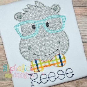 Puede incluir: Diseño bordado de un hipopótamo gris con gafas azules y una pajarita amarilla y azul. El nombre "Reese" está bordado debajo del hipopótamo.