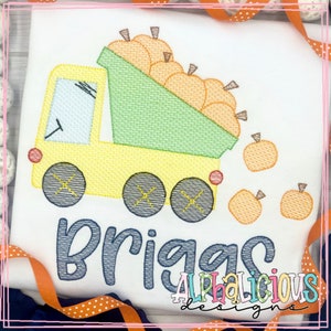 Pode incluir: Camiseta branca com um desenho bordado de um caminhão basculante amarelo cheio de abóboras laranja. O nome "Briggs" é bordado em azul abaixo. O texto "ALPHALICIOUS designs" também é visível.