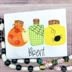 Può includere: Tre barattoli a tema Halloween con diversi design. Il primo barattolo è arancione con pois bianchi e una ragnatela. Il secondo barattolo è verde con una ragnatela bianca e un motivo a scacchi. Il terzo barattolo è giallo con un ragno nero. Il nome "Brent" è ricamato sotto i barattoli.