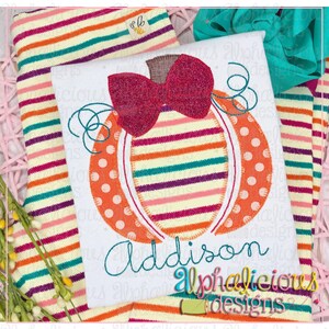 May include: White t-shirt with a colourful pumpkin design. The pumpkin features a red glitter bow, orange polka dots, and striped sections in various colours. The name "Addison" is embroidered in teal. The text "Alphalicious designs" is also visible.