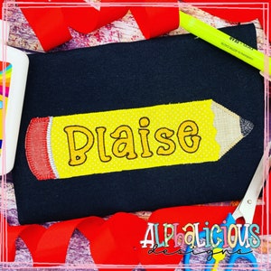May include: A yellow pencil with a red eraser and the name "Plaise" embroidered on it. The pencil is on a black fabric background.
