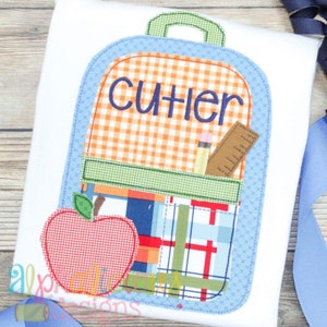 May include: A white t-shirt featuring a colorful backpack design. The backpack has a blue outline, orange gingham top, and a pocket with a pencil, ruler, and apple. The name "CUTLER" is embroidered on the backpack.