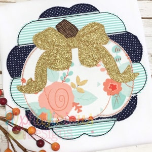 May include: A white pumpkin with a gold glitter bow, surrounded by a scalloped blue and green frame. The pumpkin is on a floral fabric background.
