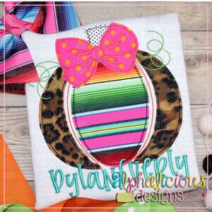 May include: A white t-shirt featuring an embroidered design of a pinata with a pink bow. The pinata is decorated with a striped pattern and a leopard print. The text "Dylan" and "alphalicious designs" is embroidered in green at the bottom of the shirt.