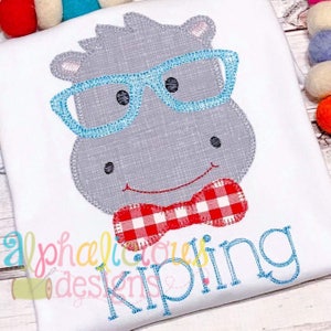 Puede incluir: Diseño bordado de un hipopótamo gris con gafas azules y una pajarita a cuadros rojos y blancos. El nombre "Kipling" está bordado debajo del hipopótamo.