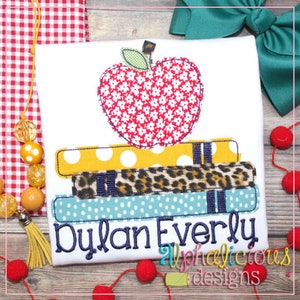 May include: A white shirt featuring a red apple with white polka dots, perched atop a stack of books with various patterns. The books are yellow with white polka dots, brown with leopard print, and blue with white polka dots. The shirt is personalised with the name "Dylan Everly" and the text "alphadelicious designs".