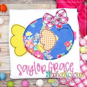 May include: A blue and yellow applique of a fish with a pink and white plaid bow on top. The text "Saylor Grace" is embroidered in pink below the fish. The text "alphalicious designs" is embroidered in pink below the name.