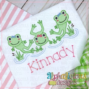 May include: Embroidered design of three green frogs with pink and blue bows, sitting on lily pads in a pond. The name "Kinnady" is embroidered below the frogs.