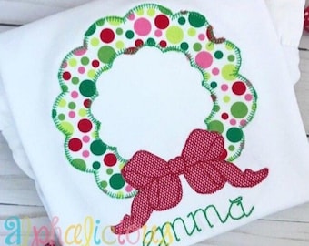 Op de afbeelding: Een wit shirt met een groene en rode kerstkrans met een rode strik en de naam "Emma" geborduurd.
