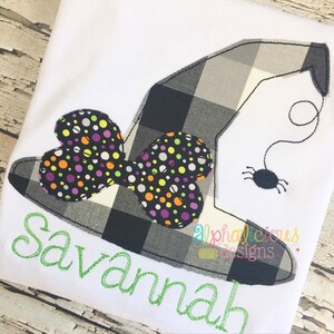 May include: White t-shirt with a witch hat design. The hat is black and grey checked with a bow featuring colourful polka dots. A small spider hangs from a thread. The name "Savannah" is embroidered in green.