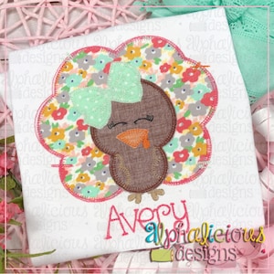 Puede incluir: Una camiseta blanca con un pavo bordado en marrón y verde sobre un fondo con estampado floral y un lazo rosa. El texto "Avery" está bordado debajo del pavo y el texto "alphalicious designs" está bordado debajo del nombre.