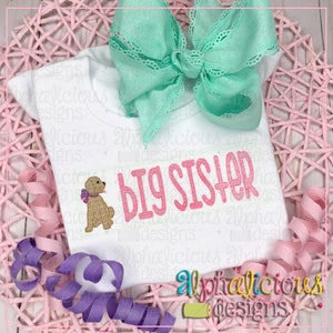 Pode incluir: Camiseta branca com um texto rosa "BIG SISTER" e um cão castanho com um laço rosa. A camiseta está sobre um fundo tecido rosa e branco com fitas rosa e roxas.