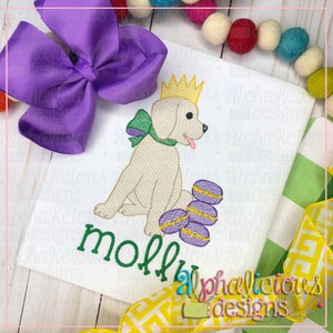 Peut inclure: Broderie représentant un chien portant une couronne et un nœud papillon vert, assis à côté d'une pile de gâteaux des rois violets et dorés. Le chien est blanc et le motif est sur un tissu blanc. Le nom "molly" est brodé sous le chien.