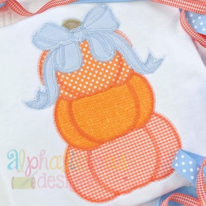 Puede incluir: Una pila de tres calabazas naranjas con un lazo azul en la parte superior. Las calabazas están decoradas con lunares blancos y cuadros naranja y blanco.