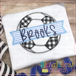 Puede incluir: Una camiseta blanca con un diseño de balón de fútbol a cuadros en blanco y negro y una pancarta a rayas azul y blanca con el nombre "BROOKS" bordado.