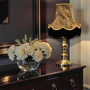 May include: A gold table lamp with a leopard print lampshade and black fringe. The lamp is on a dark wood dresser with gold handles, next to a glass bowl of white flowers. A framed artwork is in the background. The room has a classic style.