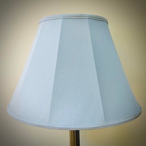 May include: A pale blue lampshade with a geometric design. The lampshade has a tapered shape and is made of a textured fabric. The top and bottom edges are trimmed with a light-coloured binding. The lampshade is mounted on a brass-coloured lamp base.