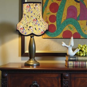 May include: A table lamp with a lampshade featuring a print inspired by Gustav Klimt's artwork. The yellow lampshade has a black scalloped edge. The lamp is on a dark brown wooden cabinet. The cabinet is in a room with artwork.