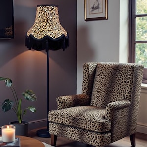 May include: A floor lamp with a leopard print lampshade and black base stands beside a matching armchair. The room includes a round wooden coffee table, a potted plant, and a framed picture. The lamp's shade has a black fringe.