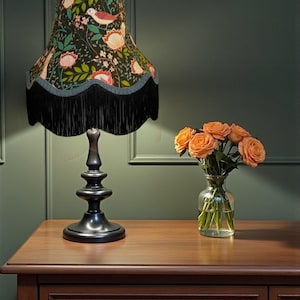 May include: A black table lamp with a floral patterned lampshade featuring birds, pink flowers, and green leaves. The lampshade has a black fringe trim. The lamp sits on a wooden table next to a vase of orange roses.