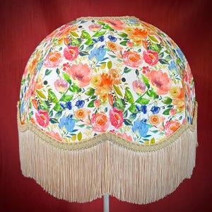 May include: A white lampshade with a floral pattern in shades of pink, orange, blue, and green. The lampshade has a scalloped edge and a fringe trim.