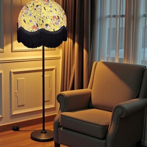 May include: A floor lamp with a round lampshade featuring a colourful, abstract design. The lampshade is trimmed with a black fringe. The lamp has a black metal pole and base. An upholstered armchair sits nearby.