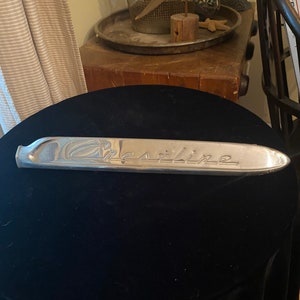 Könnte beinhalten: Ein silbernes Auto-Emblem aus Metall mit dem Wort "Gasoline" in Prägung.