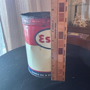 May include: Vintage metal Esso oil can with red, white, and blue colors. The can has the Esso logo and the text "IT WEIGHT" and "HUMBLE OIL & RE". A wooden ruler is placed next to the can, showing measurements in inches.