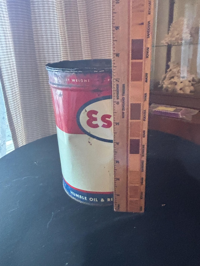 May include: Vintage metal Esso oil can with red, white, and blue colors. The can has the Esso logo and the text "IT WEIGHT" and "HUMBLE OIL & RE". A wooden ruler is placed next to the can, showing measurements in inches.
