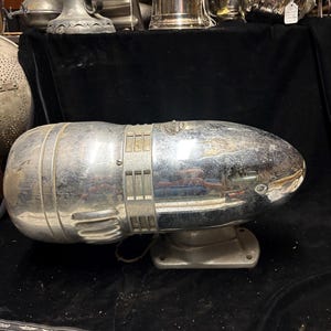 Puede incluir: Una sirena vintage cromada con una carcasa en forma de bala, que presenta ranuras de ventilación horizontales y una base de montaje central. La superficie metálica refleja la luz y un pequeño emblema es visible en la parte superior redondeada.