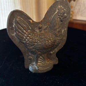 Puede incluir: Un molde de metal vintage con forma de gallo. El molde es de metal oscuro y envejecido con manchas de óxido visibles. El gallo está detallado con plumas y una cresta. El molde se utiliza probablemente para hacer chocolates o caramelos.