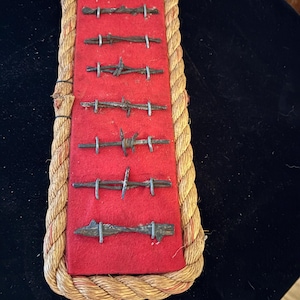 Könnte beinhalten: Eine Präsentation von sieben antiken Stacheldrahtmustern, die auf einem roten Filzhintergrund montiert sind. Die Muster sind vertikal angeordnet und mit einem dicken, gedrehten Seilrand umrahmt. Der Stacheldraht ist aus dunklem, gealtertem Metall.