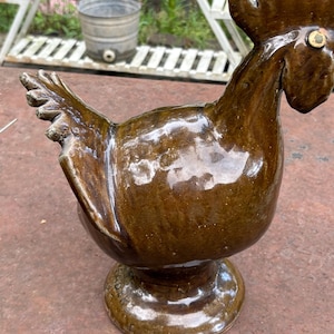 May include: A brown ceramic rooster figurine with a glossy finish. The rooster has a detailed comb, wattle, and tail feathers. It is mounted on a pedestal base. The figurine is likely handmade and suitable for home decor.