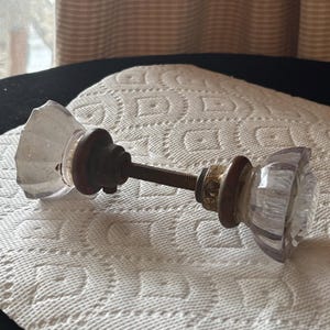 Puede incluir: Un pomo de puerta de cristal vintage con un diseño facetado. El pomo tiene una apariencia clara, similar al cristal y está unido a una varilla de metal oscuro con detalles decorativos de latón. El pomo es probablemente para un armario o una puerta.