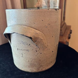 May include: A stoneware crock with a handle, featuring the text "J.D. Craven" and the number "1". The crock has a light brown color with a slightly textured surface and a dark rim. It is an antique kitchenware item.