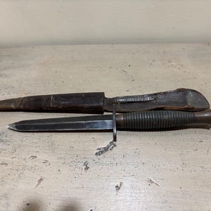 Puede incluir: Un cuchillo vintage con una hoja oscura y un mango acanalado, acompañado de una funda de cuero. La hoja del cuchillo es puntiaguda y el mango tiene un diseño texturizado y estriado. La funda es marrón y muestra signos de desgaste.